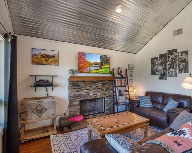 A cozy living room with a brick fireplace, a wooden coffee table, leather sofas, and artwork on the walls, featuring a television mounted above the fireplace.