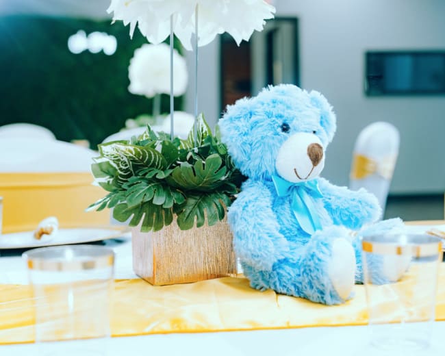 A blue teddy bear is seated next to a floral centerpiece on a table set for an event.