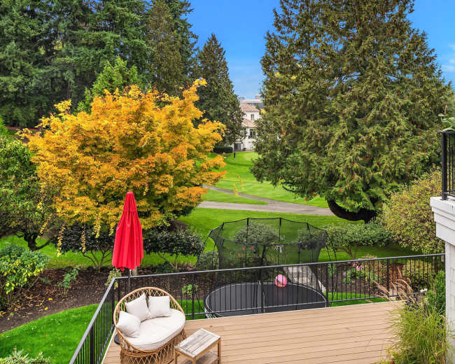 A backyard scene featuring a deck with seating, a bright red umbrella, and a view of a manicured green lawn bordered by trees and shrubs.