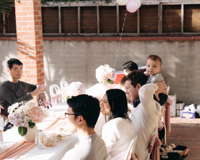 A group of people sits at a long table decorated with flowers, while one person holds a baby on their shoulder.