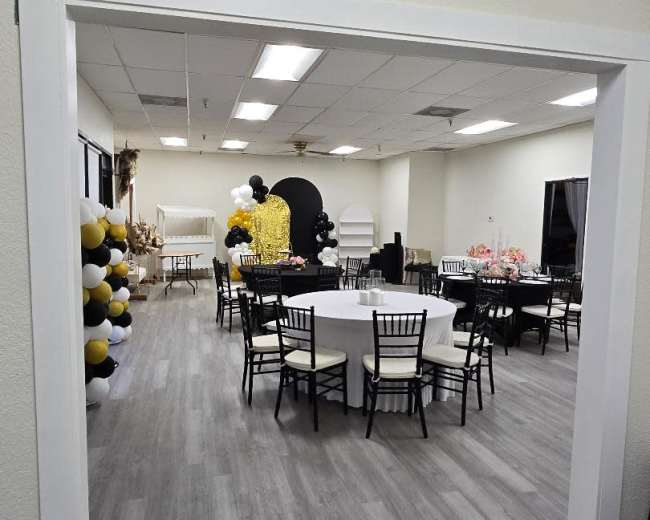 The image shows a spacious event room with round tables set for guests, black and gold balloon decorations, and a presentation area at the far end.