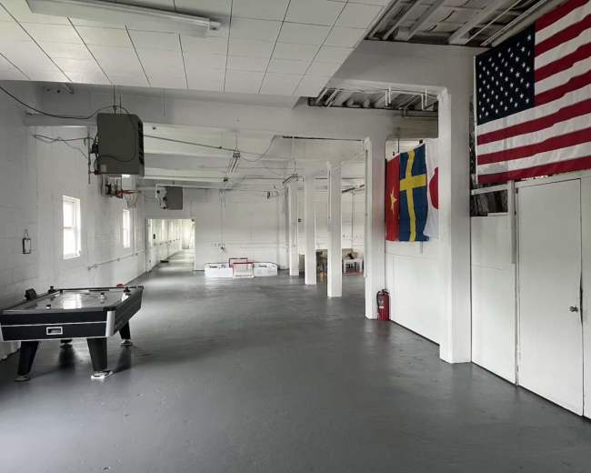 The image shows a large, empty indoor space with a pool table, several flags on the walls, and a white painted floor.