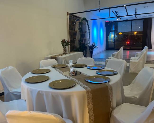 A round table with eight gold plates is set for a meal in a dimly lit dining area that features white draped chairs and blue lighting.
