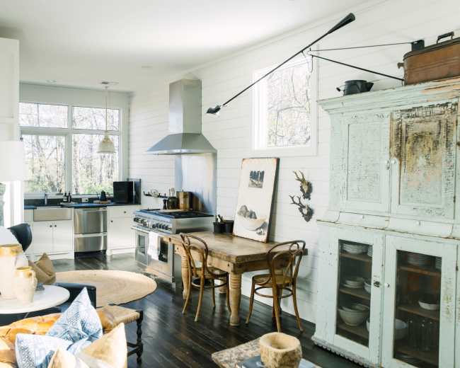 The image shows a modern kitchen with stainless steel appliances, a wooden dining table, and a light-filled living area featuring a cozy seating arrangement and decorative items.