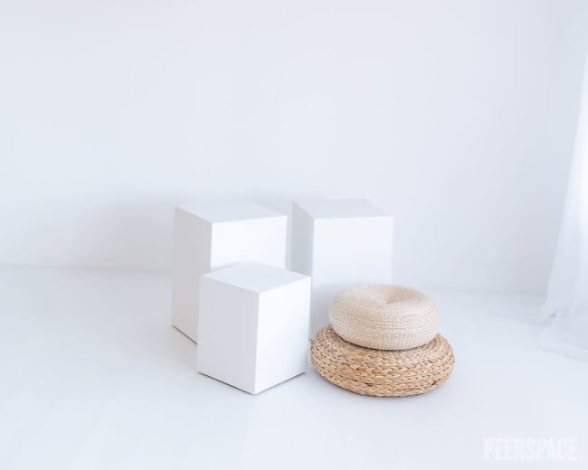 The image features three white cube-shaped objects and a round woven hat resting on a light-colored floor in a minimalist space with a white backdrop.