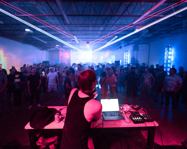 A DJ performs on stage in front of an audience, with vibrant lasers illuminating the space around them.
