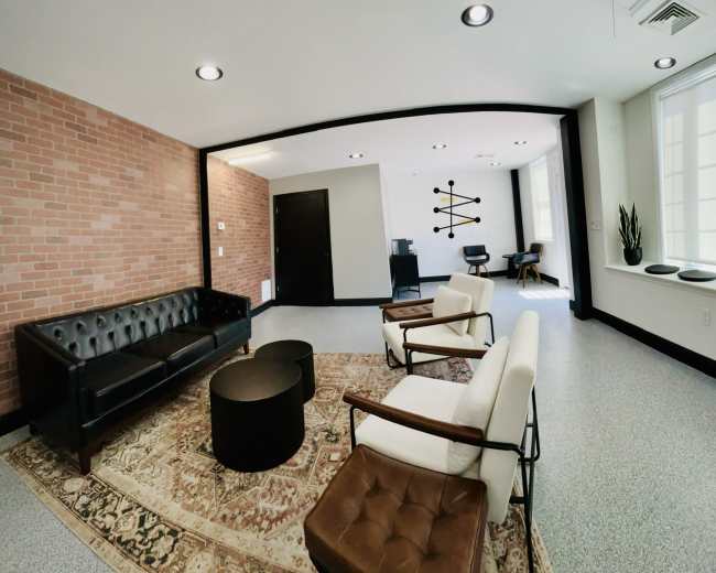 The image shows a modern lounge area featuring a black leather sofa, two white armchairs, a round black coffee table, and a patterned rug on a speckled floor.