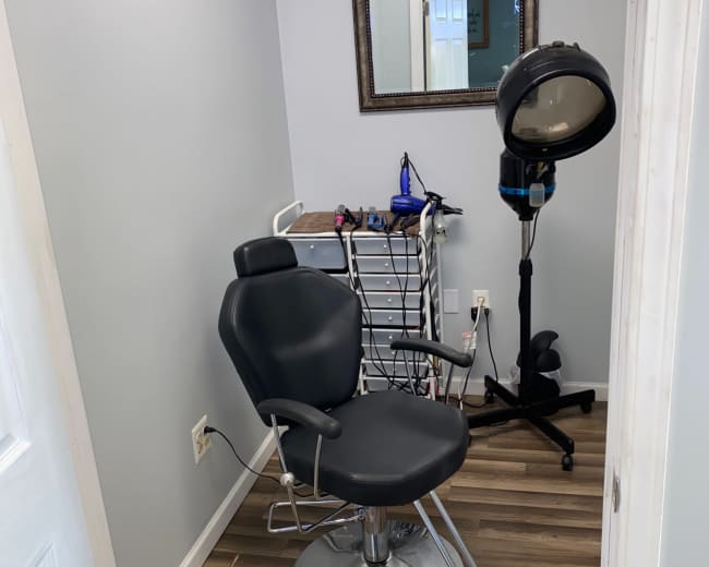 The image shows a hair salon corner featuring a black salon chair, a mirror on the wall, and a hooded hairdryer stand beside a rack with various hair styling tools.