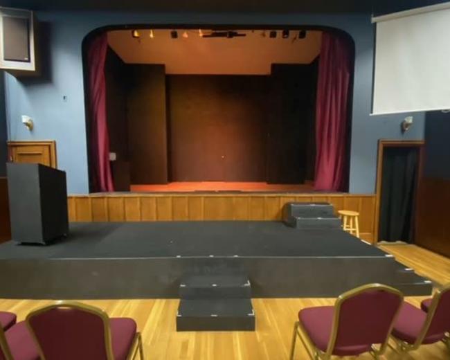 A small stage in a theatre, framed by maroon curtains, with a podium and several chairs arranged in front.