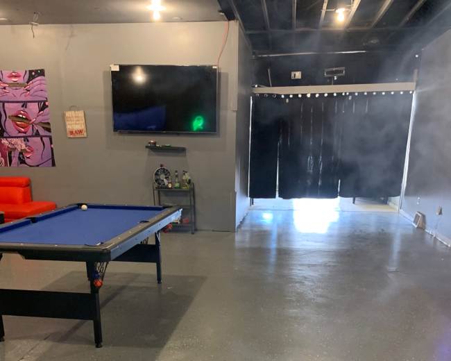 The image shows a room with a polished concrete floor, a blue pool table in the foreground, a large television mounted on the wall, and a doorway with black curtains in the background.