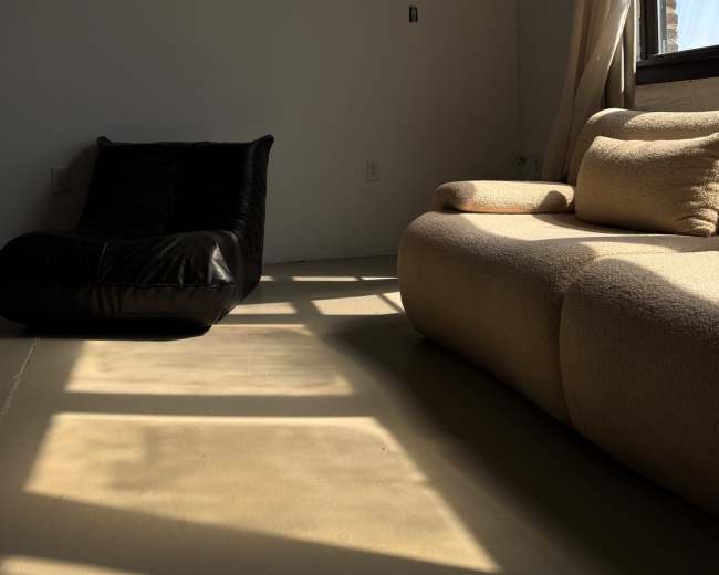 The image shows a minimalist interior with a black bean bag chair positioned next to a cream-colored couch, illuminated by sunlight streaming through a large window.