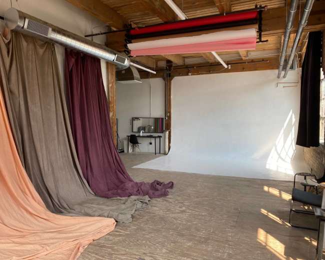 The image shows a photography studio with draped backdrops in various colors and a plain white wall, along with a wooden floor and a desk setup.