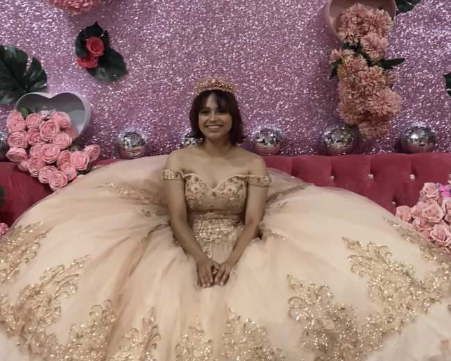 A girl in a golden embellished dress sits on a pink sofa surrounded by a sparkling pink backdrop adorned with flowers.