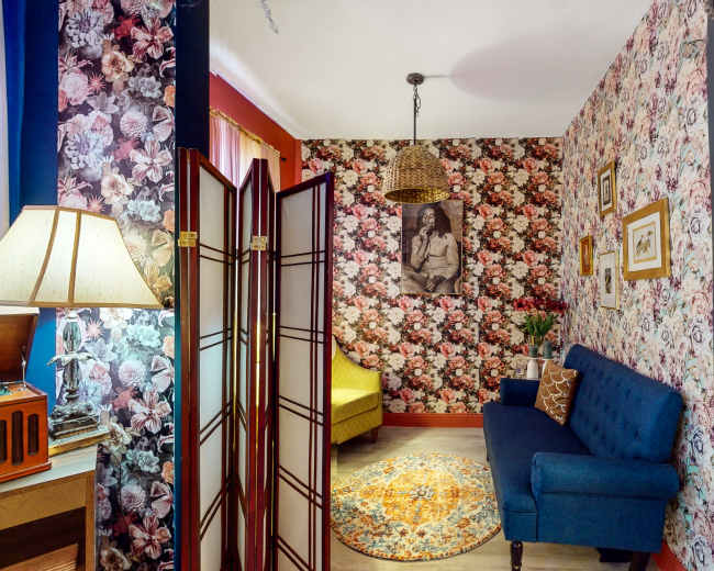 A cozy room with floral wallpaper featuring a blue sofa, a lamp, a screen divider, and a round rug, complementing a vintage radio in the corner.