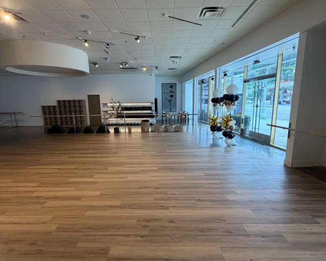 The image shows a spacious, empty dance studio with wooden flooring and large windows allowing natural light to enter.
