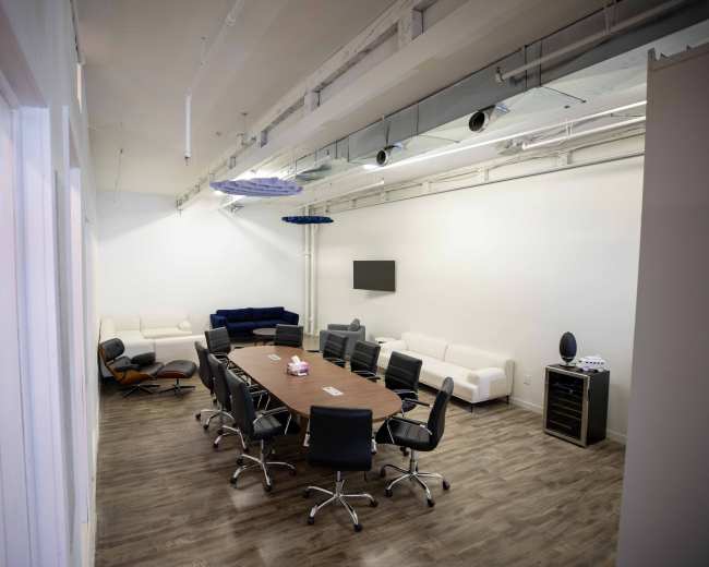 The image shows a modern conference room with a large oval table surrounded by black chairs, a couch in the corner, and a wall-mounted screen.