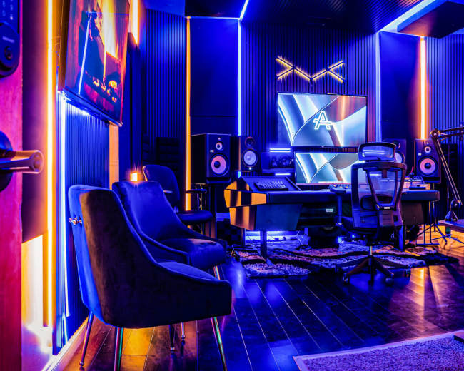 A modern recording studio features a mixing desk, speakers, and colorful LED lighting, with seating and acoustic treatment on the walls.