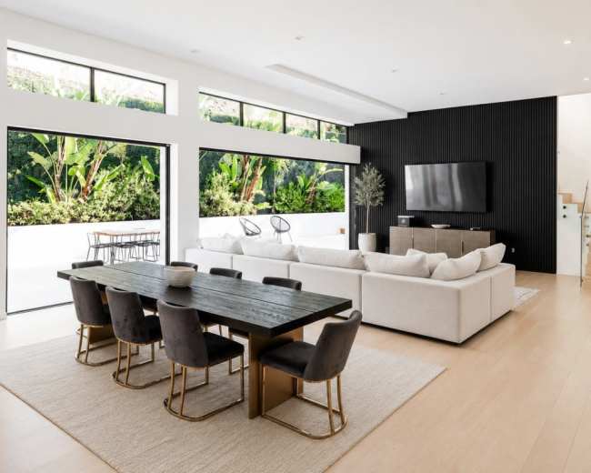 The image shows a modern living and dining area with a large sectional sofa, a dining table set for eight, and large windows overlooking a green outdoor space.