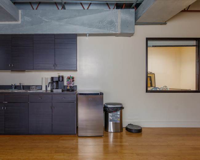 The image shows a small kitchen area with dark cabinets, a sink, a coffee machine, a stainless steel refrigerator, and a trash can, alongside a window looking into another room.