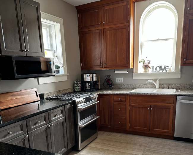 The image shows a kitchen featuring dark and light wooden cabinets, stainless steel appliances, and a countertop with a sink beneath a window.