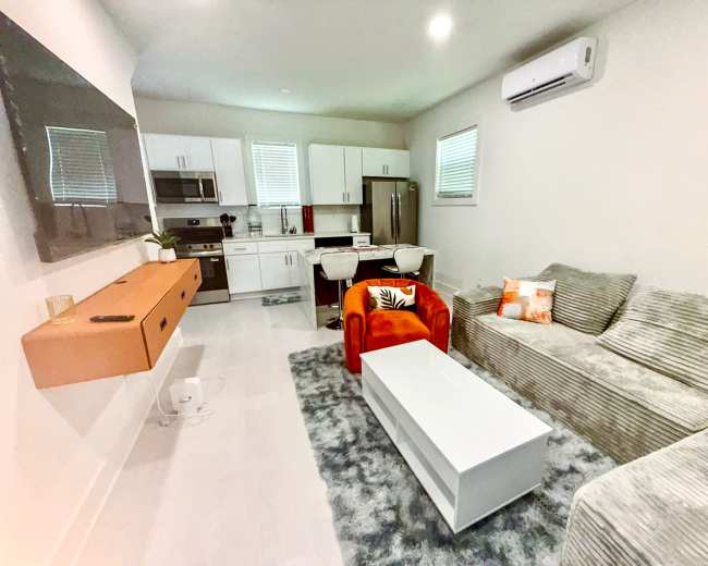 The image shows a modern living room with a gray sofa, an orange chair, a white coffee table, and a kitchen area in the background.