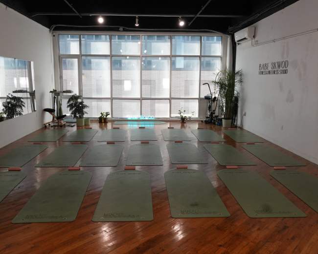 The image shows a spacious yoga studio with neatly arranged mats on a wooden floor and large windows letting in natural light.