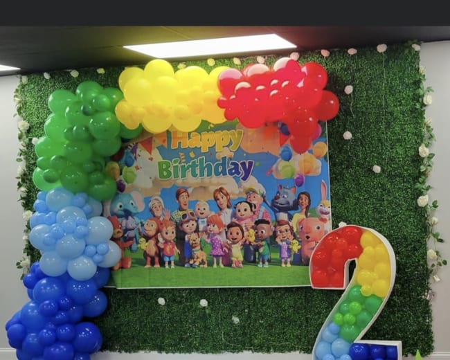 A birthday celebration setup featuring colorful balloon arrangements and a "Happy Birthday" sign against a green backdrop.