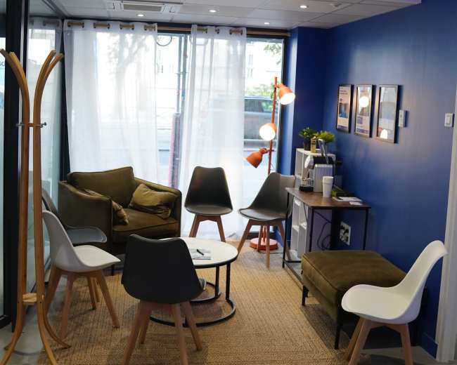 The image depicts a modern lounge area with a mix of seating, including chairs and sofas, arranged around a central table on a woven rug in front of a blue wall.