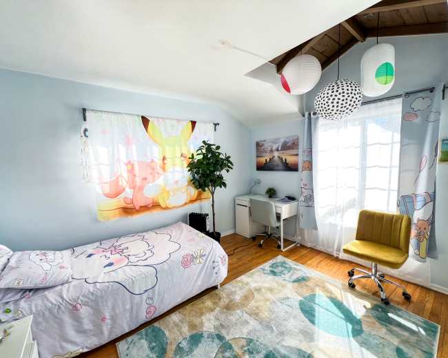 The image shows a brightly decorated bedroom with a bed featuring a playful bedspread, a desk and chair in one corner, and a window adorned with colorful paper lanterns.