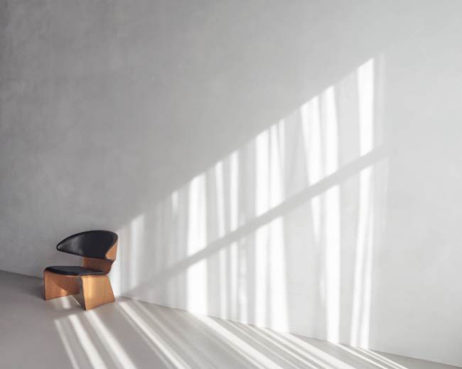 A wooden chair with a black seat is positioned against a gray wall, with shadows from vertical blinds creating stripes of light on the floor.