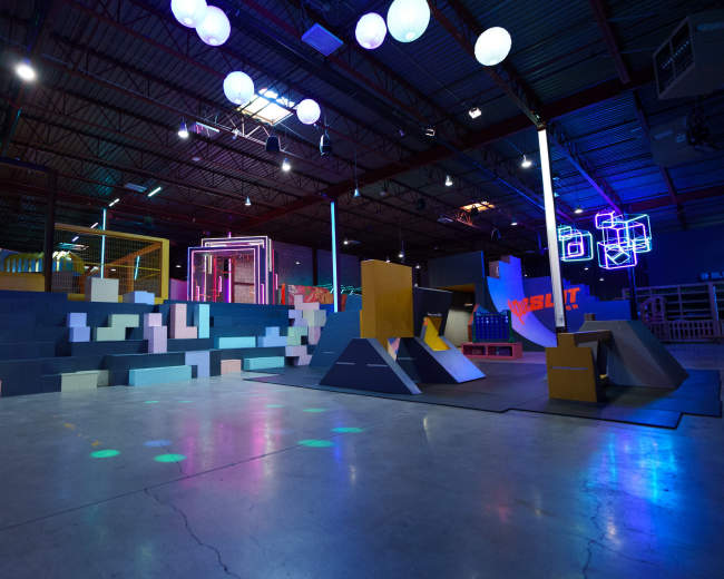 The image shows an indoor skatepark with various ramps and obstacles illuminated by colorful lights.
