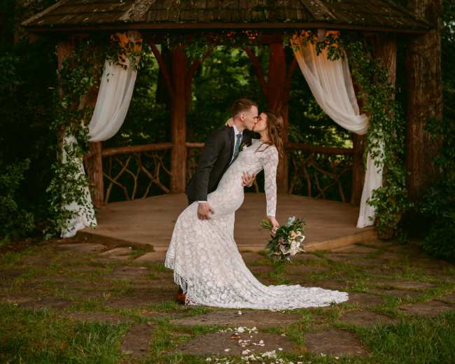 A couple in wedding attire shares a kiss under a gazebo surrounded by trees.