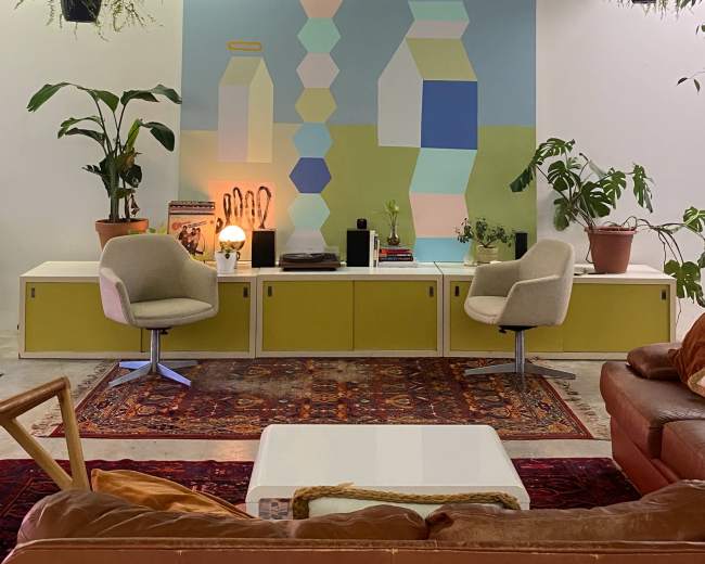 A modern living room features a mural of abstract shapes on the wall, with plants and contemporary furniture arranged around a coffee table.
