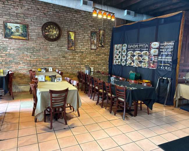 The image shows the interior of a restaurant featuring wooden tables and chairs, with a partially visible menu board and decorative brick walls.