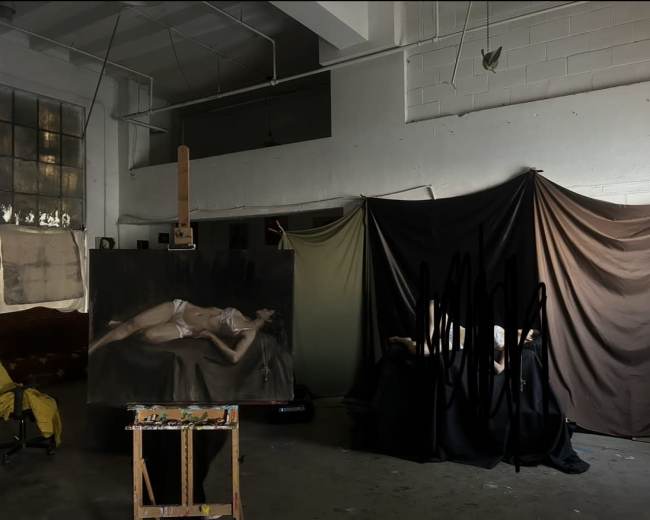 The image depicts an art studio with a partially completed painting of a reclining figure on an easel, while several draped fabrics create a backdrop on one side.