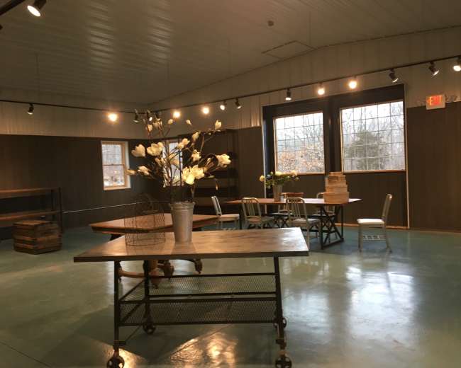 The image shows a spacious room with wooden tables, chairs, and decorative flowers, illuminated by overhead lighting and large windows.