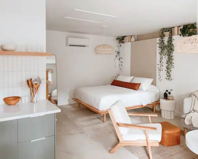 A modern, minimalist bedroom with a wooden bed, a small sitting area, and plants adorning the walls.