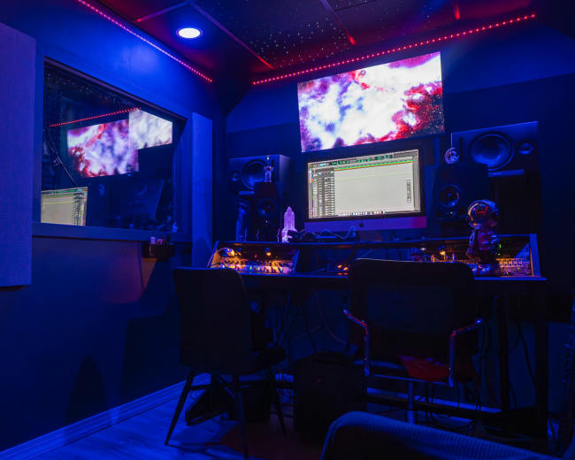 The image shows a music production studio with a large computer monitor, sound speakers, and colorful LED lights illuminating the room.