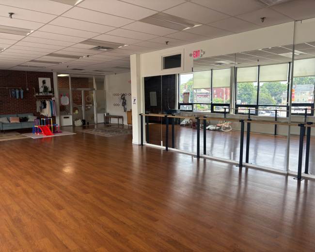The image shows an empty studio space with wooden flooring, large mirrors on one wall, and a play area with colorful equipment in the corner.