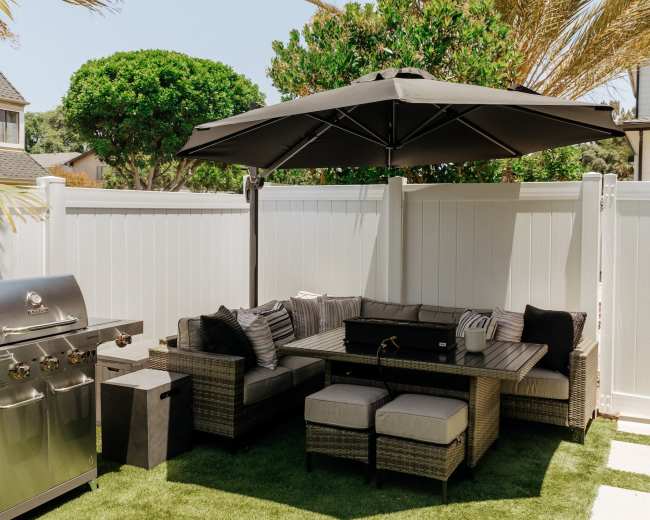 An outdoor seating area with a sectional sofa under a large umbrella, surrounded by palm trees and a grill on the side.