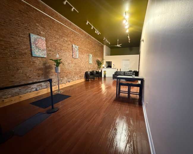 The image shows an empty, spacious interior with exposed brick walls, wooden flooring, a reception area at the back, and seating arrangements on the left side.