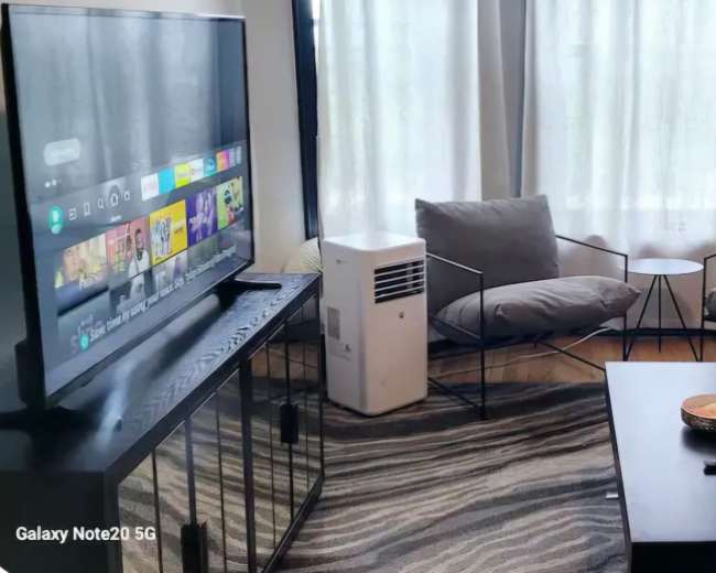 The image shows a living room with a beige sofa, two gray armchairs, a small side table, and a television on a stand, all set on a patterned rug.