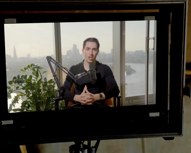 A man sits in front of a microphone and camera, speaking in a well-lit room with a city skyline visible through the large window behind him.