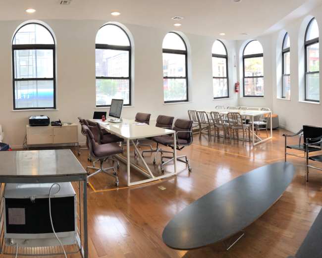 The image shows a modern, open-plan office space with large arched windows, a coffee machine, seating areas, and multiple tables arranged for work and meetings.