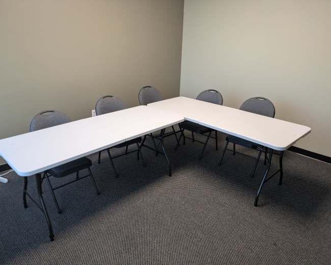 The image shows two rectangular tables arranged in a T formation, with six gray chairs placed around them in a room with beige walls and a carpeted floor.