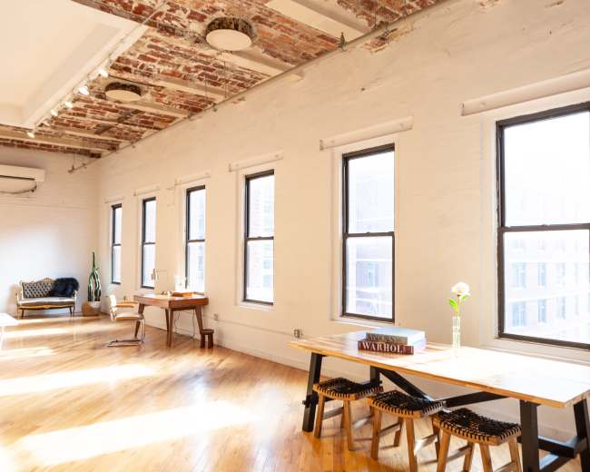 A bright, airy room features large windows, wooden floors, and minimalist furniture, including tables and chairs.