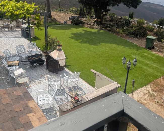 The image shows a green lawn surrounded by a stone patio, outdoor furniture, and an outdoor grill, with trees and hills in the background.