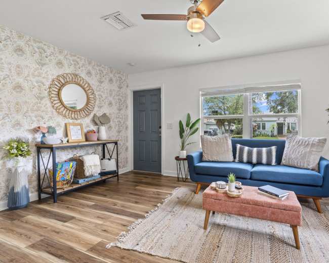 A modern living room featuring a blue sofa with patterned pillows, a coffee table, a plant in the corner, and decorative elements against a wallpapered wall.