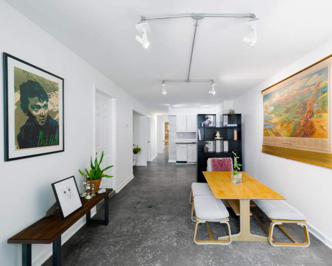 The image shows a modern dining area with a wooden table, gray upholstered chairs, artwork on the walls, and a bookshelf against one side.
