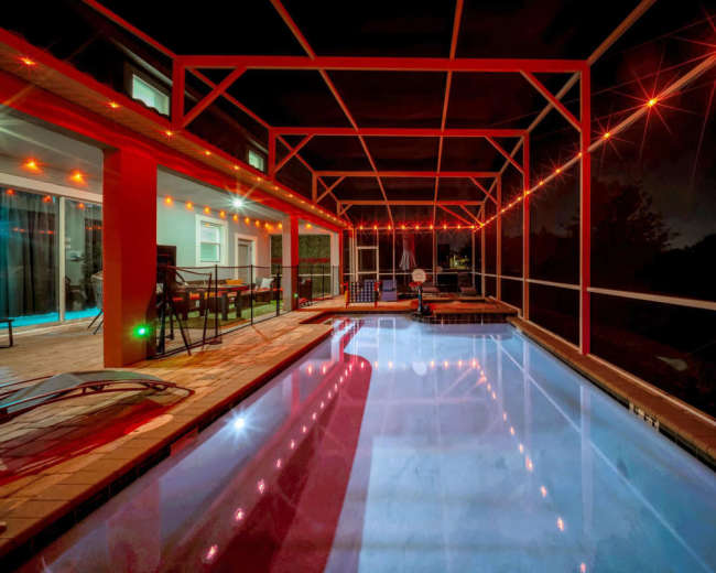 The image shows a screened-in pool area at night, illuminated by string lights and featuring a patio with chairs and a table.